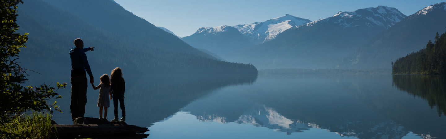 An adult stands on the shoreline with two people, admiring the view of the lake and the mountains in the distance.