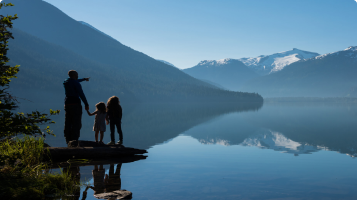 An adult stands on the shoreline with two people, admiring the view of the lake and the mountains in the distance.