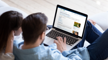 A man and a woman are sitting together. They are browsing through the Better Homes website on the laptop perched on his lap.