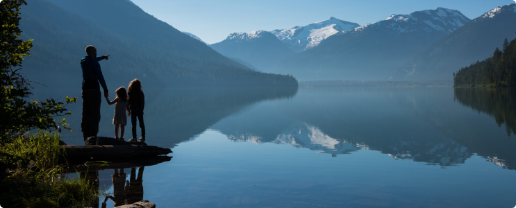 An adult stands on the shoreline with two people, admiring the view of the lake and the mountains in the distance.