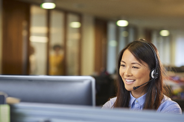 An Asian woman is working as a customer service representative. She is wearing a headset with a desktop computer in front of her.