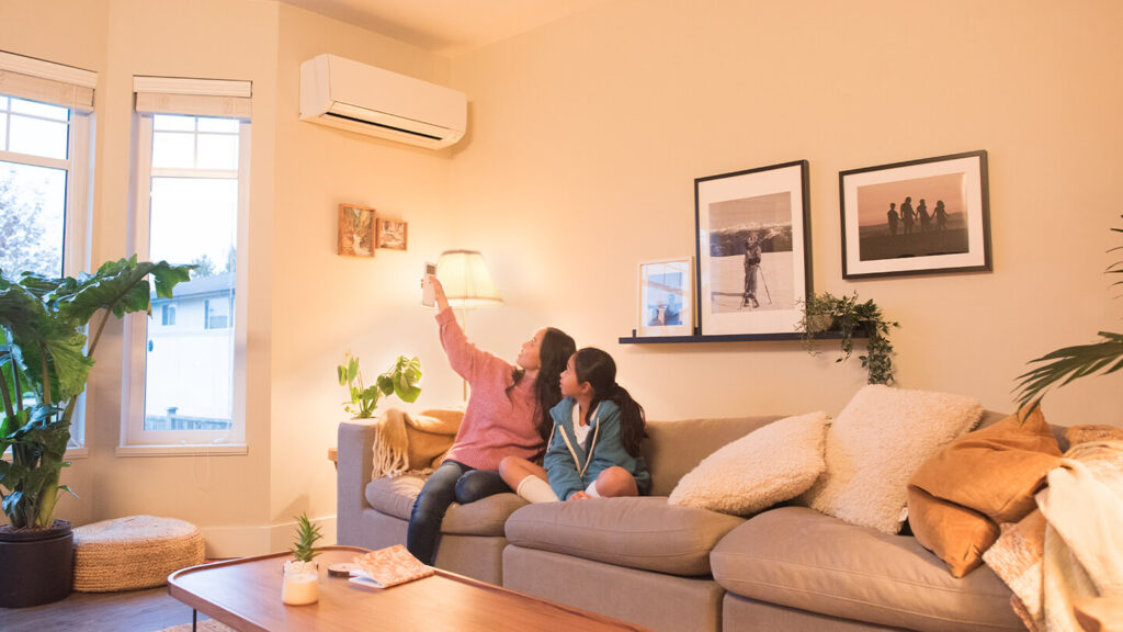 A mother and daughter sitting together on a couch in a comfortable living room pointing a remote at the indoor unit of their heat pump.
